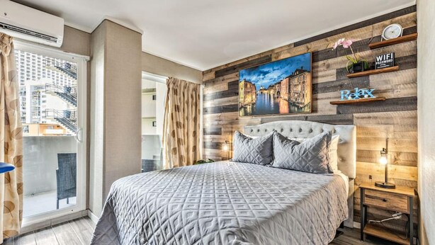 Une chambre moderne avec un grand lit, un tableau représentant un paysage urbain, un panneau mural en bois et un balcon offrant une vue sur les bâtiments.