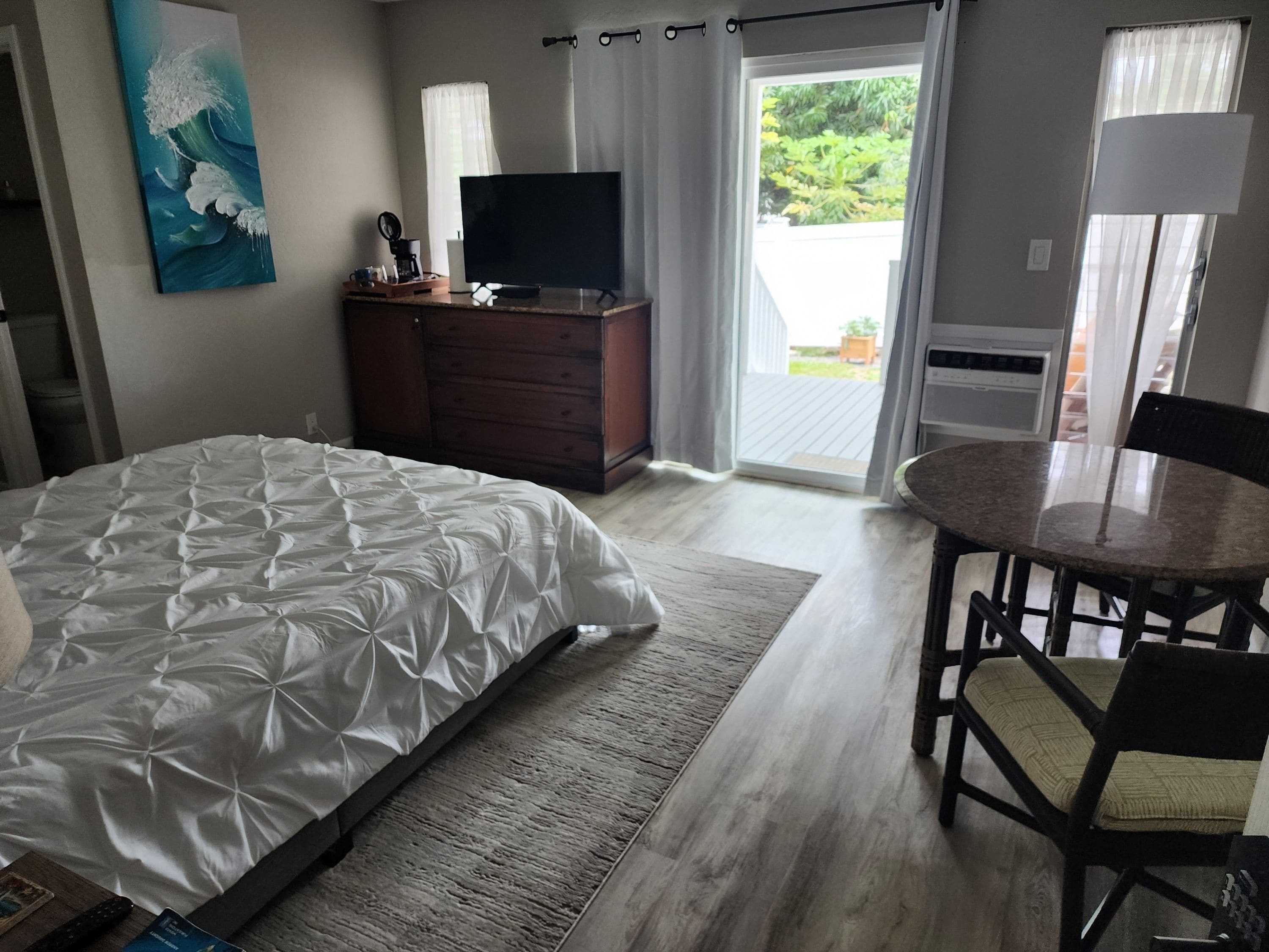 Habitación de hotel con cama, televisor, cómoda de madera, mesa de comedor con sillas y vistas al exterior.