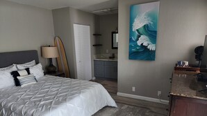 Un dormitorio con una cama grande, cabecera de madera, mesita de noche, lámpara, una tabla de surf, armario y un cuadro en la pared.