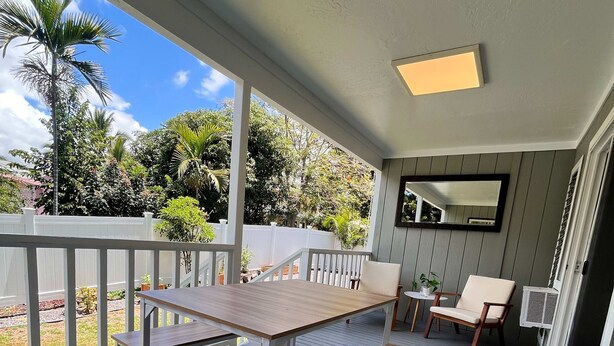 Un patio techado al aire libre con una mesa y bancos de madera, barandal blanco y vista a la vegetación.