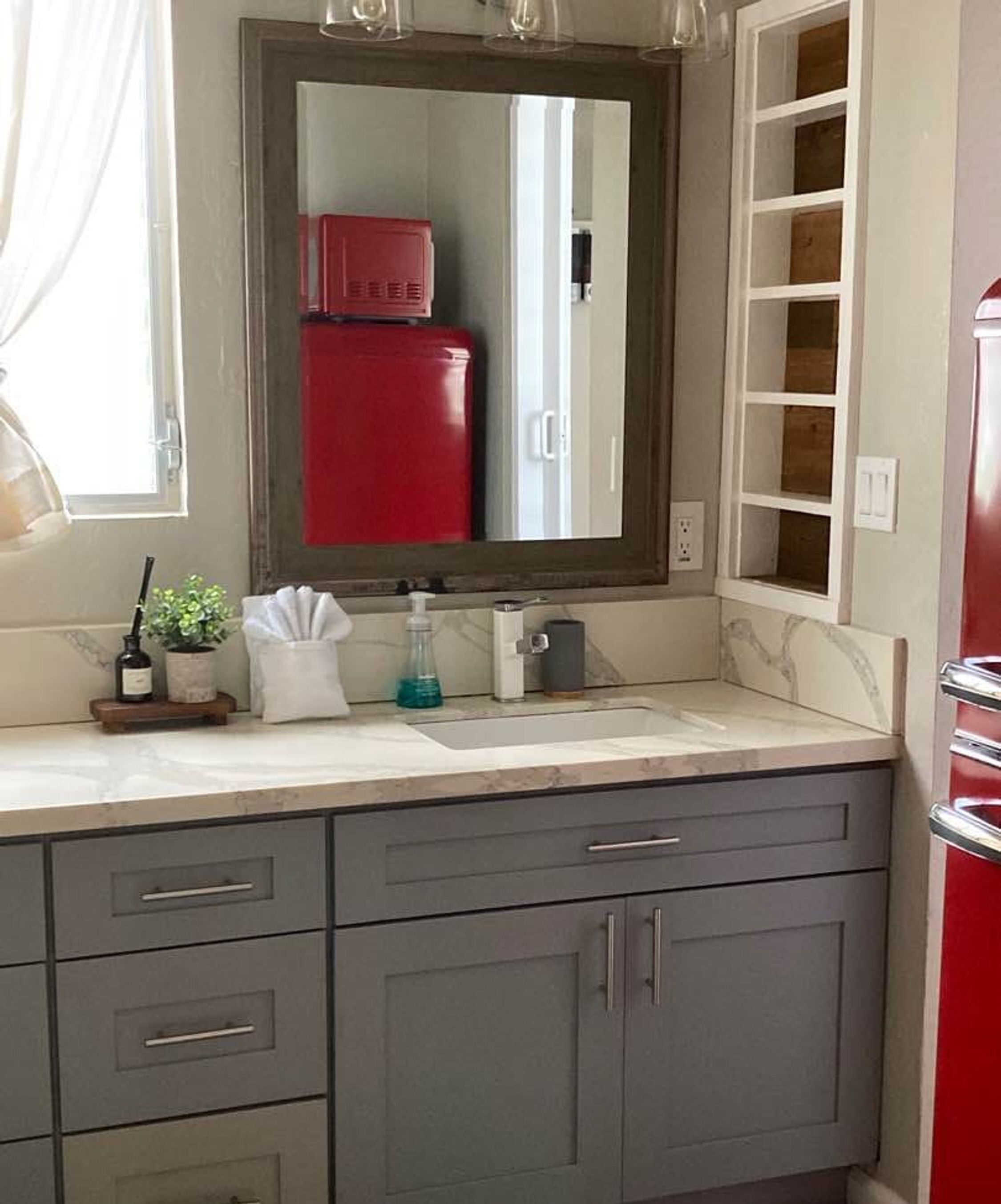 Une salle de bain avec un comptoir en marbre, un miroir, un appareil électroménager rouge et des meubles gris.