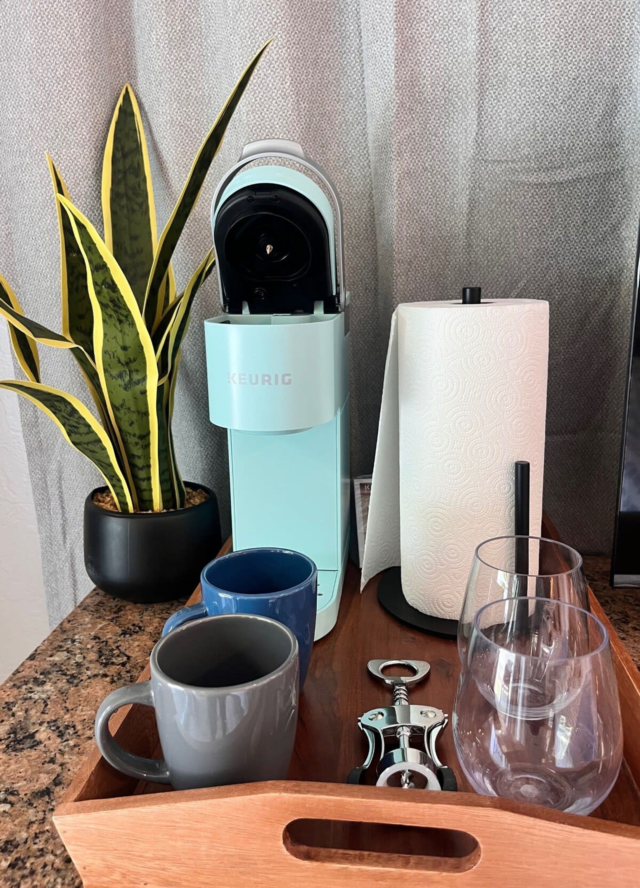 Een koffiezetapparaat, een pak tissues, een blauwe mok, een grijze mok, een wijnglas en een kurkentrekker op een houten dienblad.