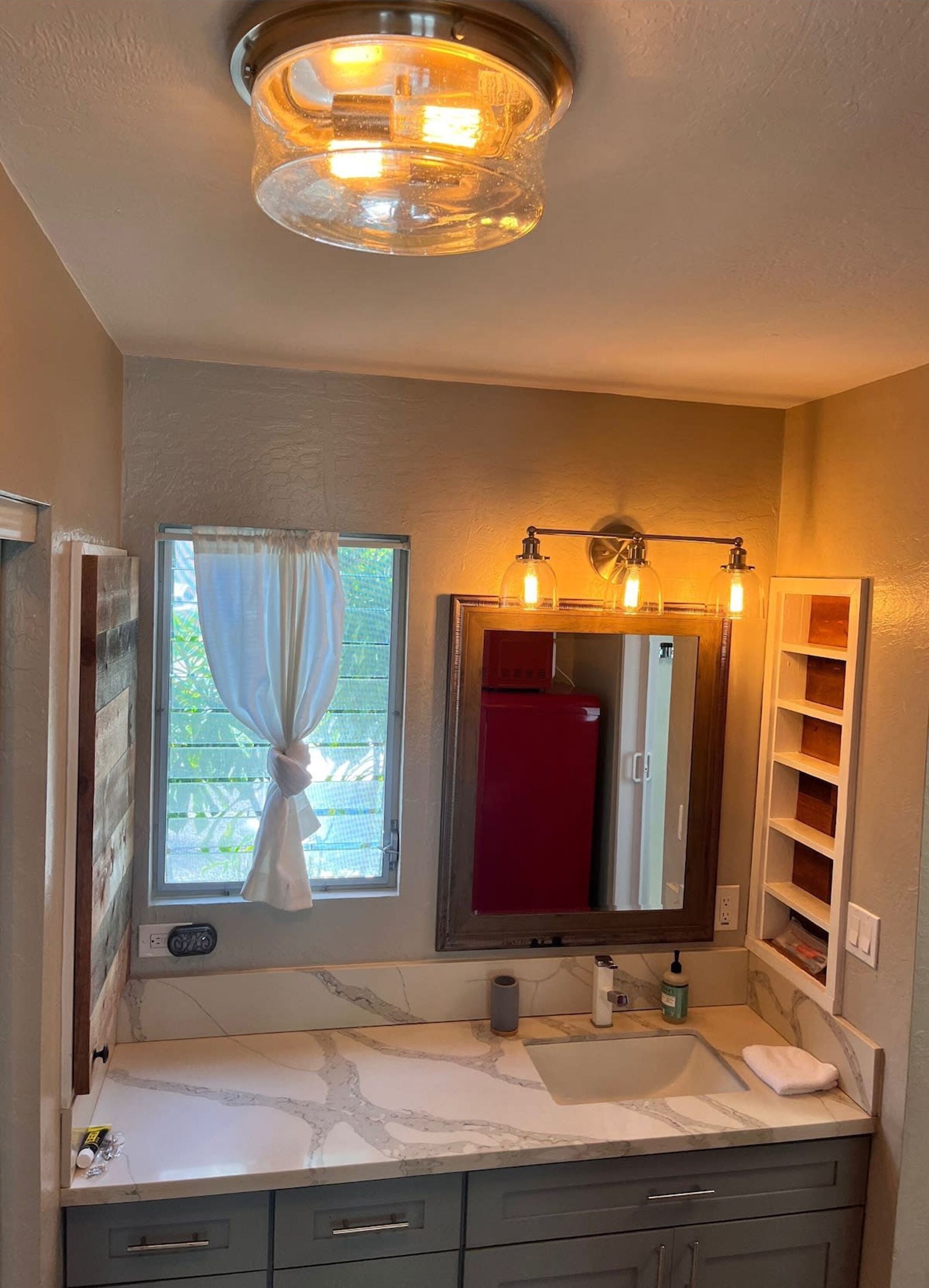 A bathroom with a marble countertop, a sink, a mirror with lights, and a window with curtains.