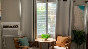 Une pièce avec une table en bois et deux chaises, une fenêtre avec des rideaux, une plante en pot et un tableau accroché au mur.