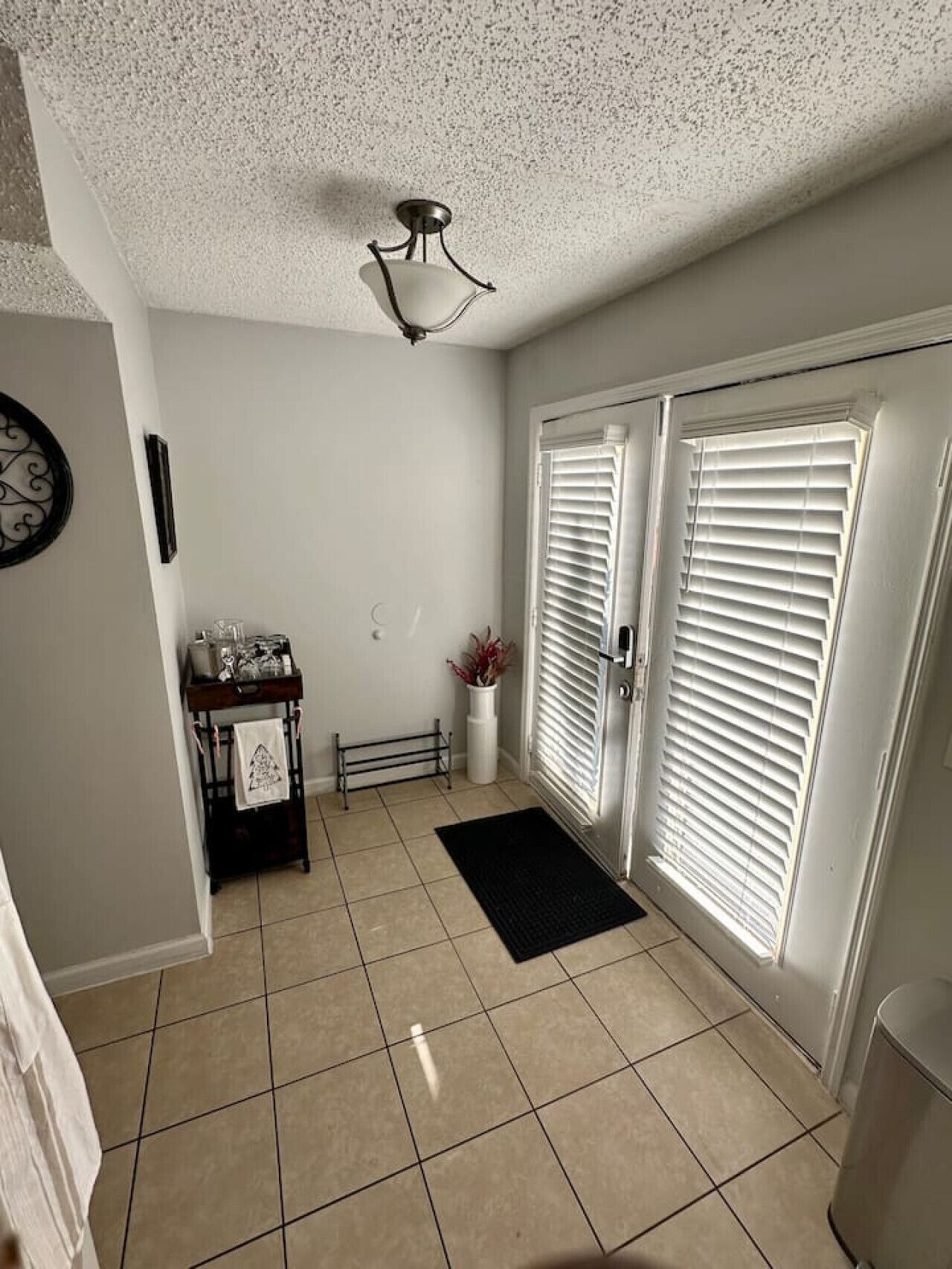 Un couloir avec un sol carrelé, un luminaire au plafond, une porte avec des stores blancs, une petite table sur laquelle se trouve un vase, et un paillasson noir à l’entrée.