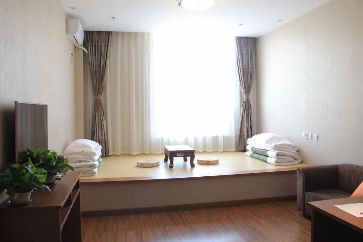 Ein Hotelzimmer mit großem Fenster, einem kleinen Tisch, einer Couch, einem Fernseher und einer Pflanze.