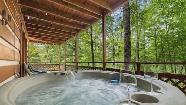 A hot tub with a wooden deck and glass railings, surrounded by a forested area.