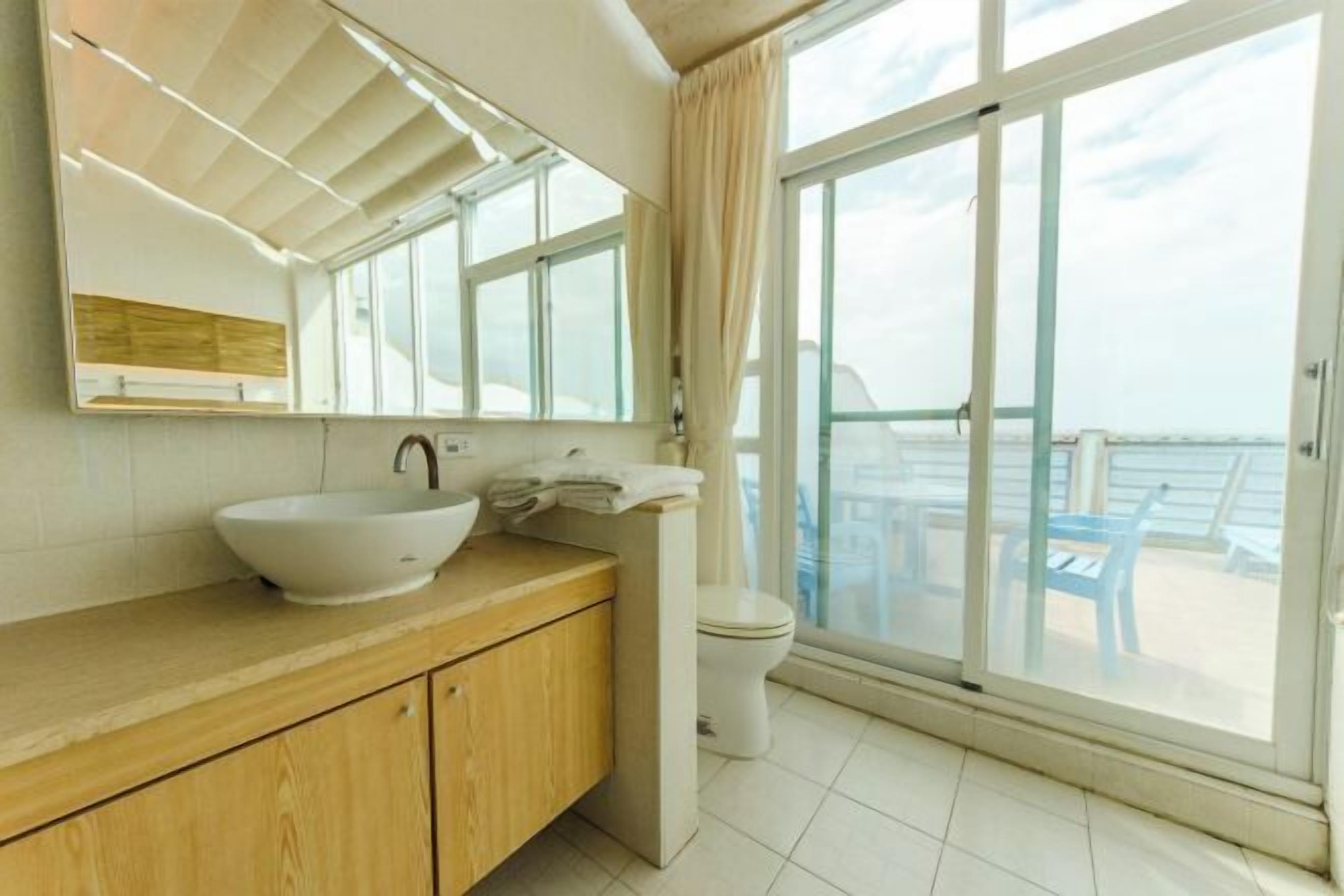 Un baño moderno con un ventanal grande, un tocador de madera, un lavabo blanco y un inodoro.
