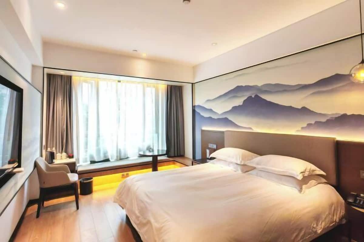 A modern hotel room with a large bed, a desk, a chair, and a window with curtains.