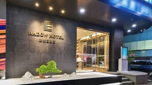 Et hotels moderne facade med en markant indgang, reception og et anlagt område med et lille træ og sten.