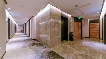 Corredor moderno de hotel com piso de mármore, paredes revestidas de madeira e um elevador.