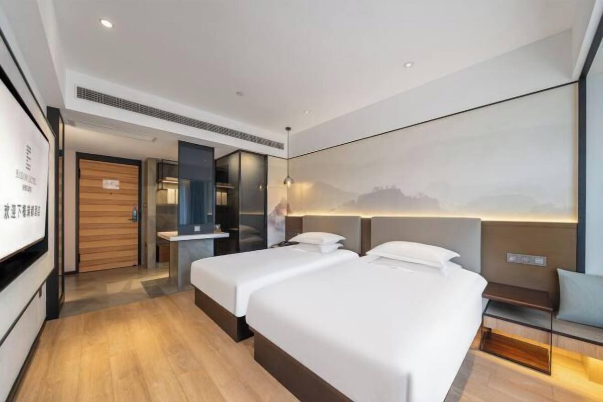 A modern hotel room with two beds, a large wall-mounted TV, and a view of a mountainous landscape.