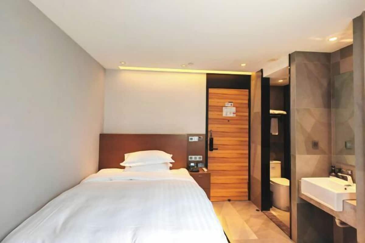 A modern hotel room with a bed, a wooden headboard, a nightstand, a bathroom with a sink and mirror, and a door leading to another room.