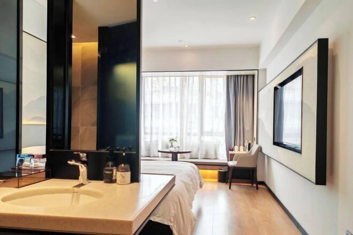 A modern hotel room with a large bathroom sink, a bed, a small table, and a window with curtains.