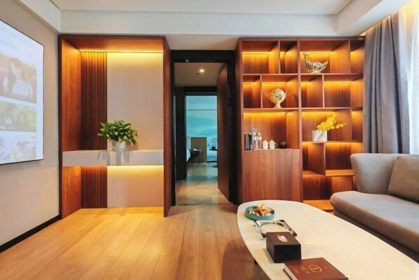 A modern hotel room with a wooden reception desk, a coffee table with a book and a remote control, a sofa, and a wall-mounted shelf with decorative items.