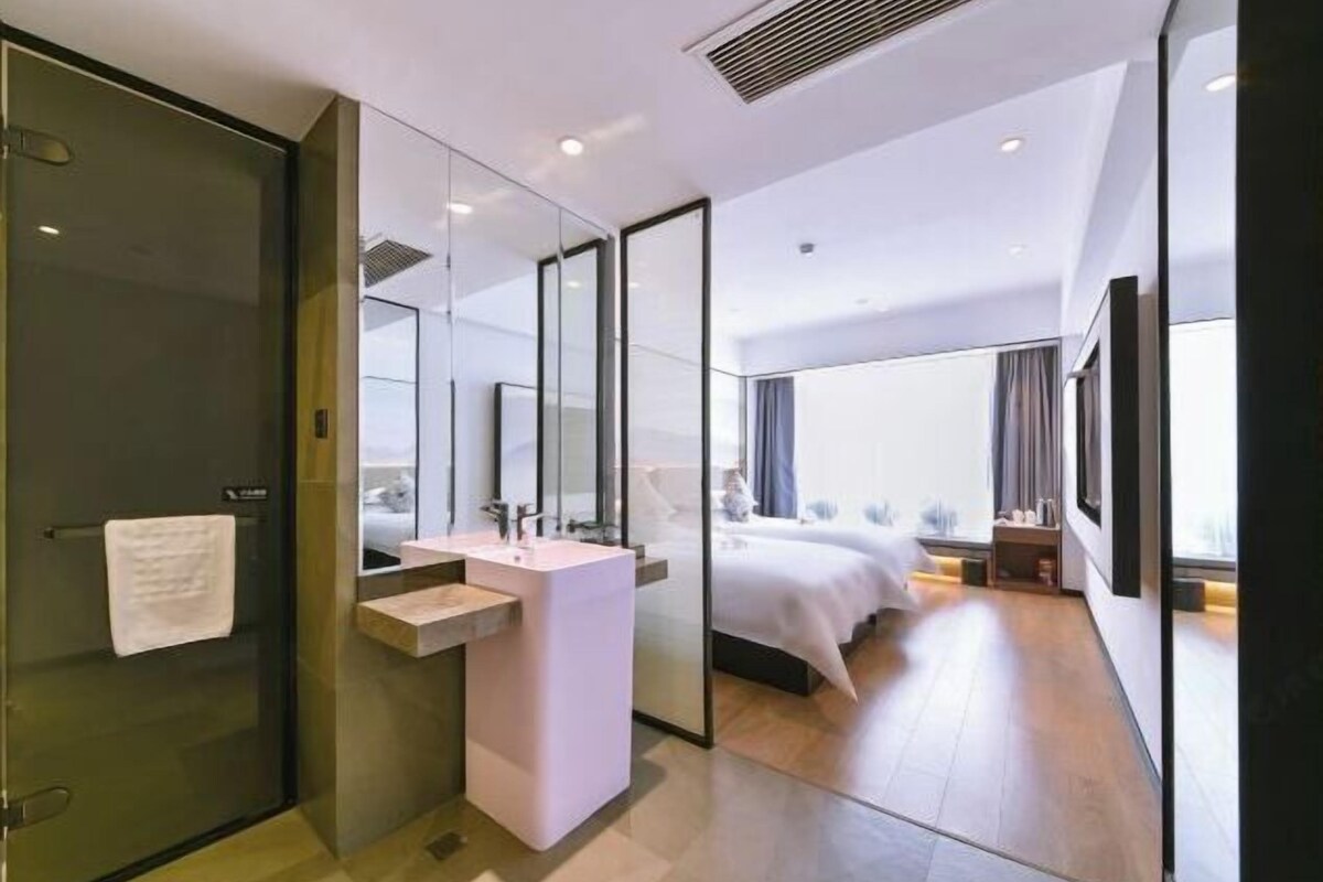 A modern hotel room with a glass shower enclosure, a white sink, a large bed, and a view of the city.
