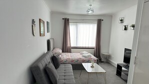 A modern, compact room with a grey sofa, a small white table, a TV, and a bed with a floral bedspread.