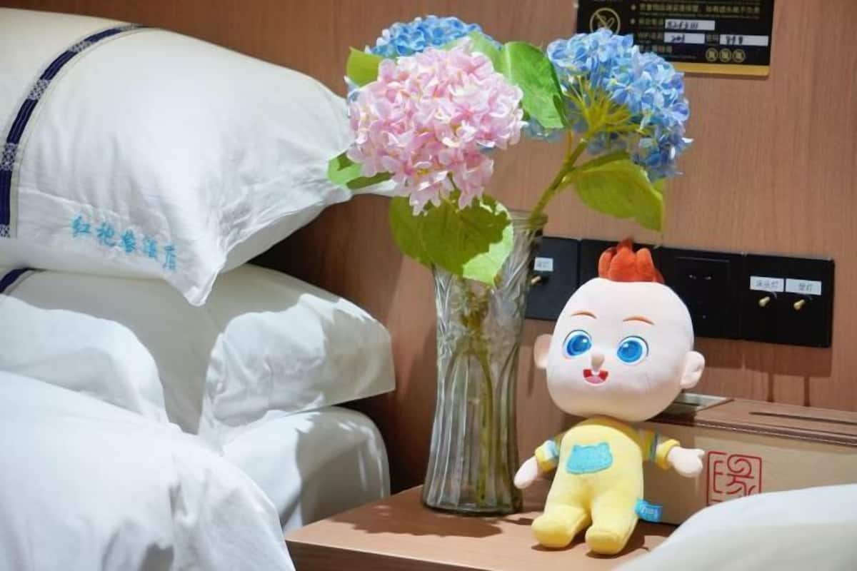 A hotel room with a bed, pillows, a vase with flowers, a plush toy, and a wall-mounted control panel.