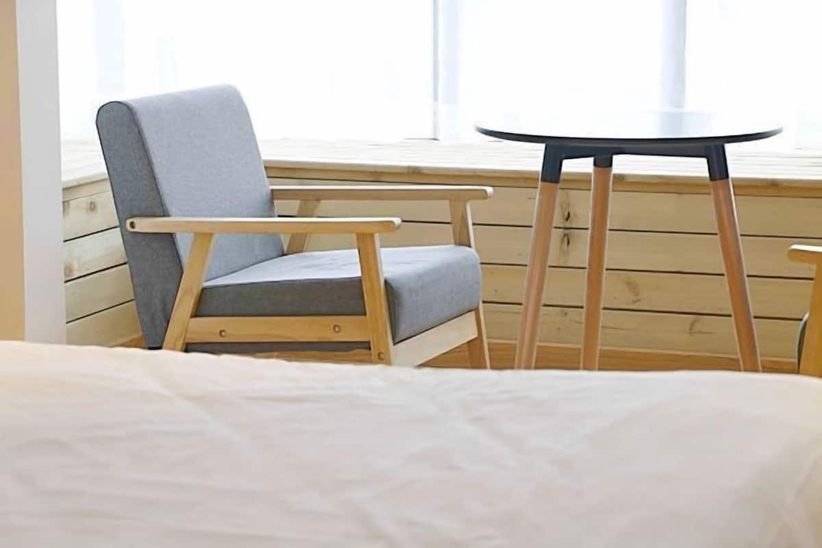 A room with a bed, a grey upholstered chair, and a small round table with wooden legs.