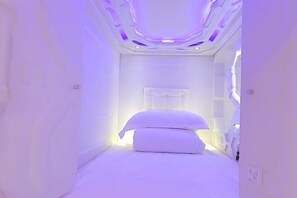 Una habitación pequeña y estrecha, de color blanco, con una cama, cabecera blanca y un ventanal.