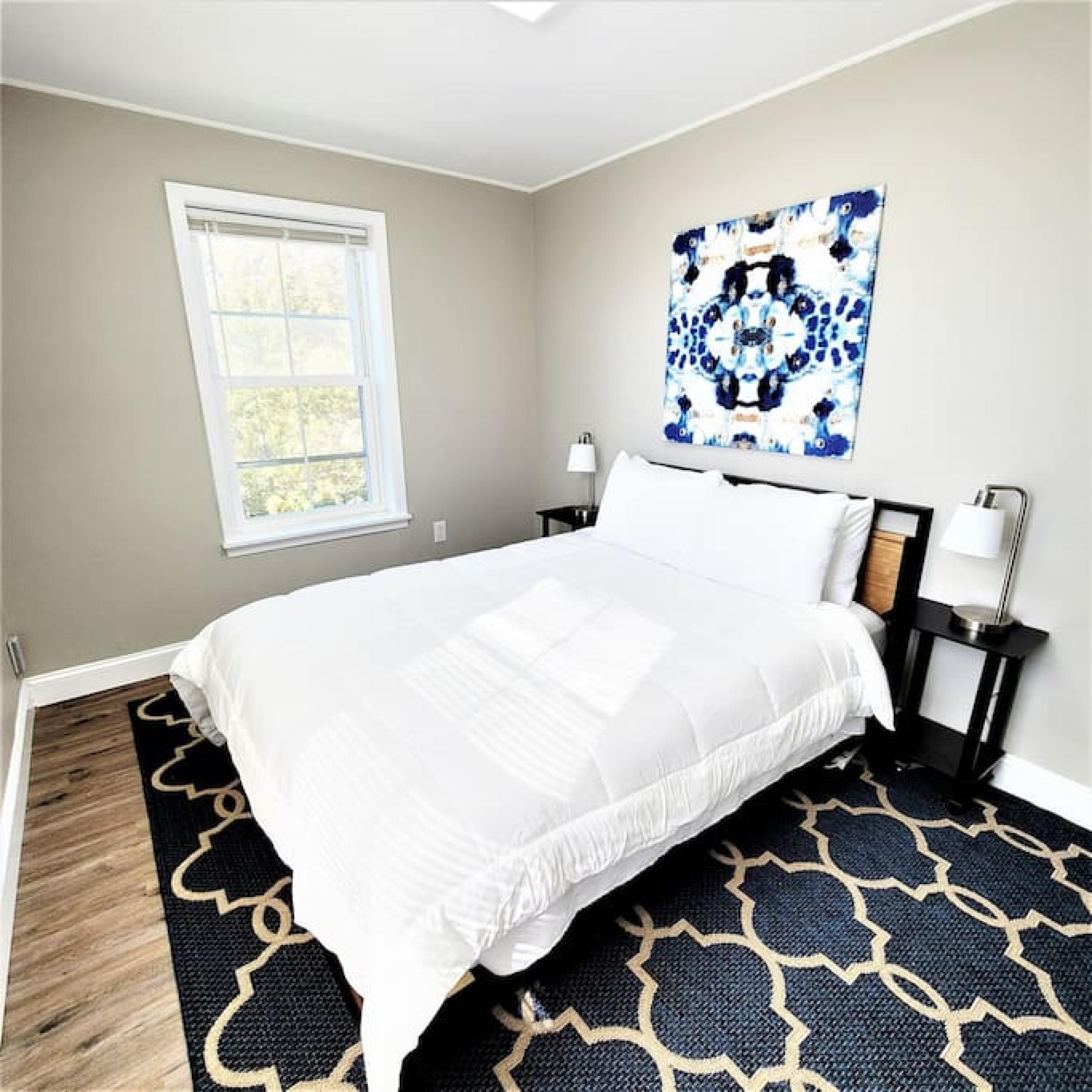 A bedroom with a bed, a window, a wall art piece, and a nightstand with a lamp.