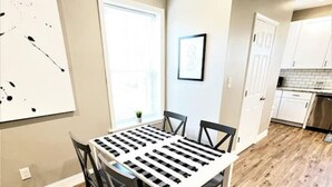 A dining area with a white table and chairs, a window with blinds, and a kitchen in the background.