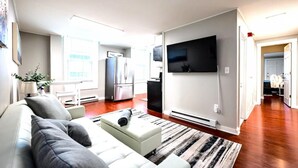 Una sala de estar moderna con un sofá blanco, un televisor de pantalla plana montado en la pared y una zona de cocina al fondo.