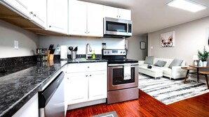 Cocina moderna con armarios blancos, encimera negra, electrodomésticos de acero inoxidable y un sofá blanco en la zona de estar.
