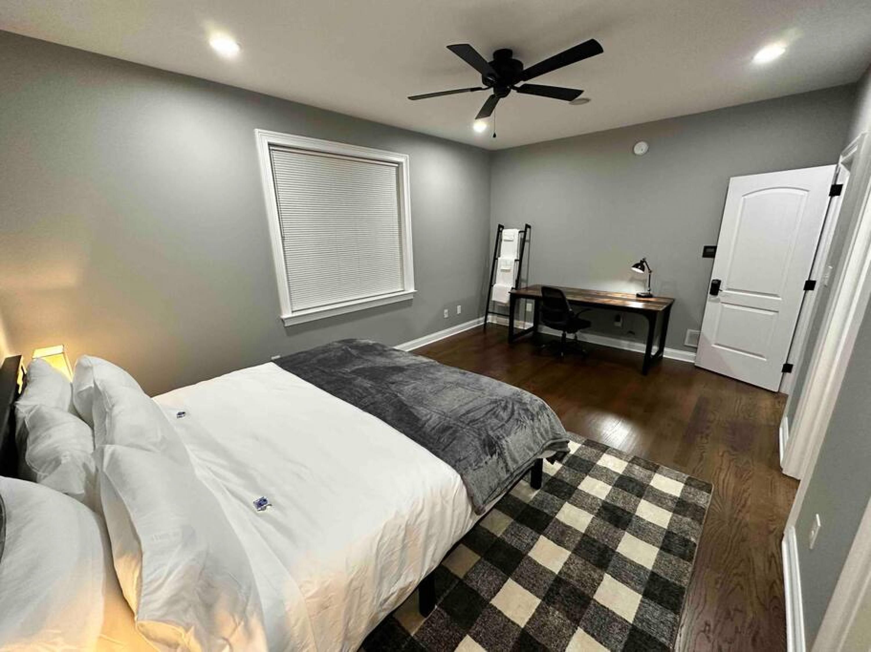 A bedroom with a bed, a desk, a chair, a ceiling fan, a window with blinds, and a door.