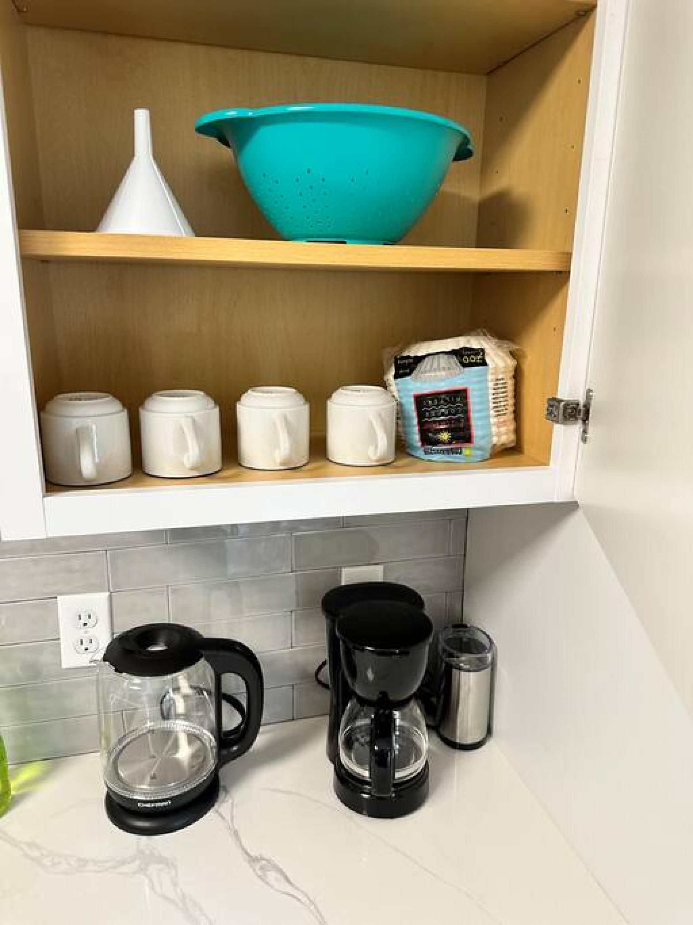 A kitchen with a wooden shelf holding a blue colander, a white strainer, and a bag of flour. Below, there are two coffee makers and a glass container.