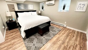 A modern bedroom with a large bed, a nightstand, a lamp, an air conditioner, and a window with blinds. - Boardwalk- Large Modern Unit With King bed Fast Wifi Free Coffee and Easy Parking (Wilmington)