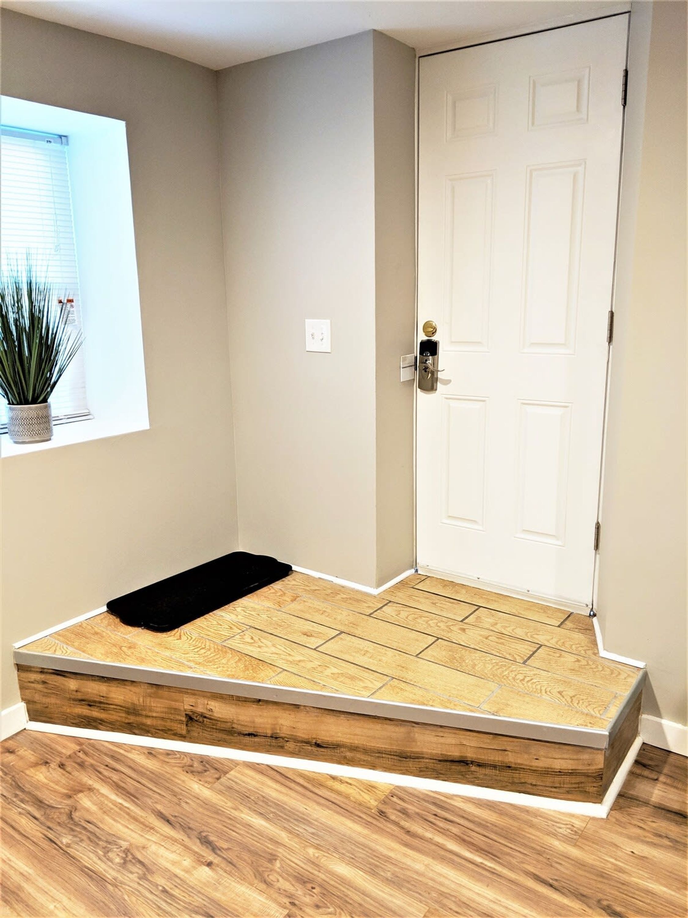 A room with a wooden floor, a white door, a small window with blinds, and a potted plant.