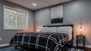 Un dormitorio con cama, mesita de noche, lámpara, ventana con persianas y una obra de arte colgada en la pared.