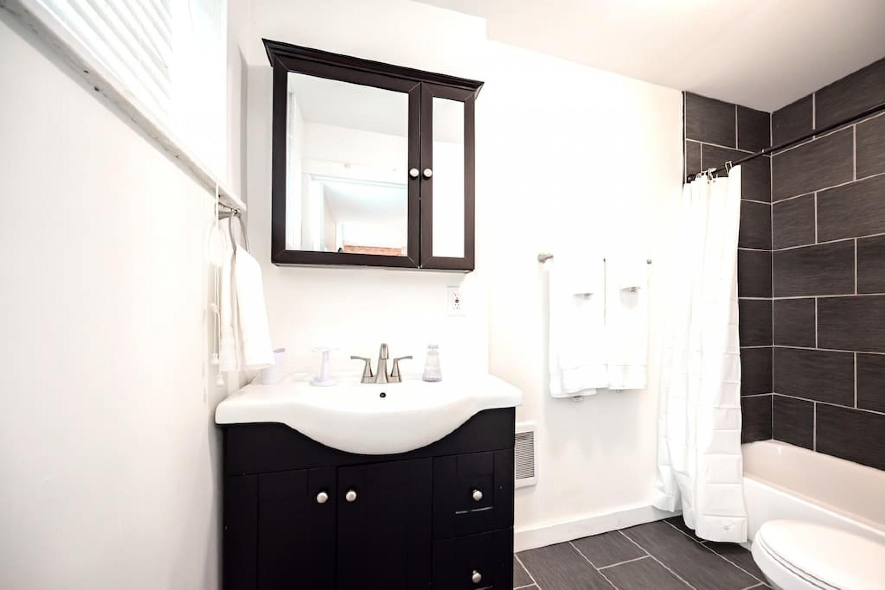Un baño moderno con un lavabo blanco, muebles de baño negros, un espejo grande y paredes cubiertas de baldosas oscuras.