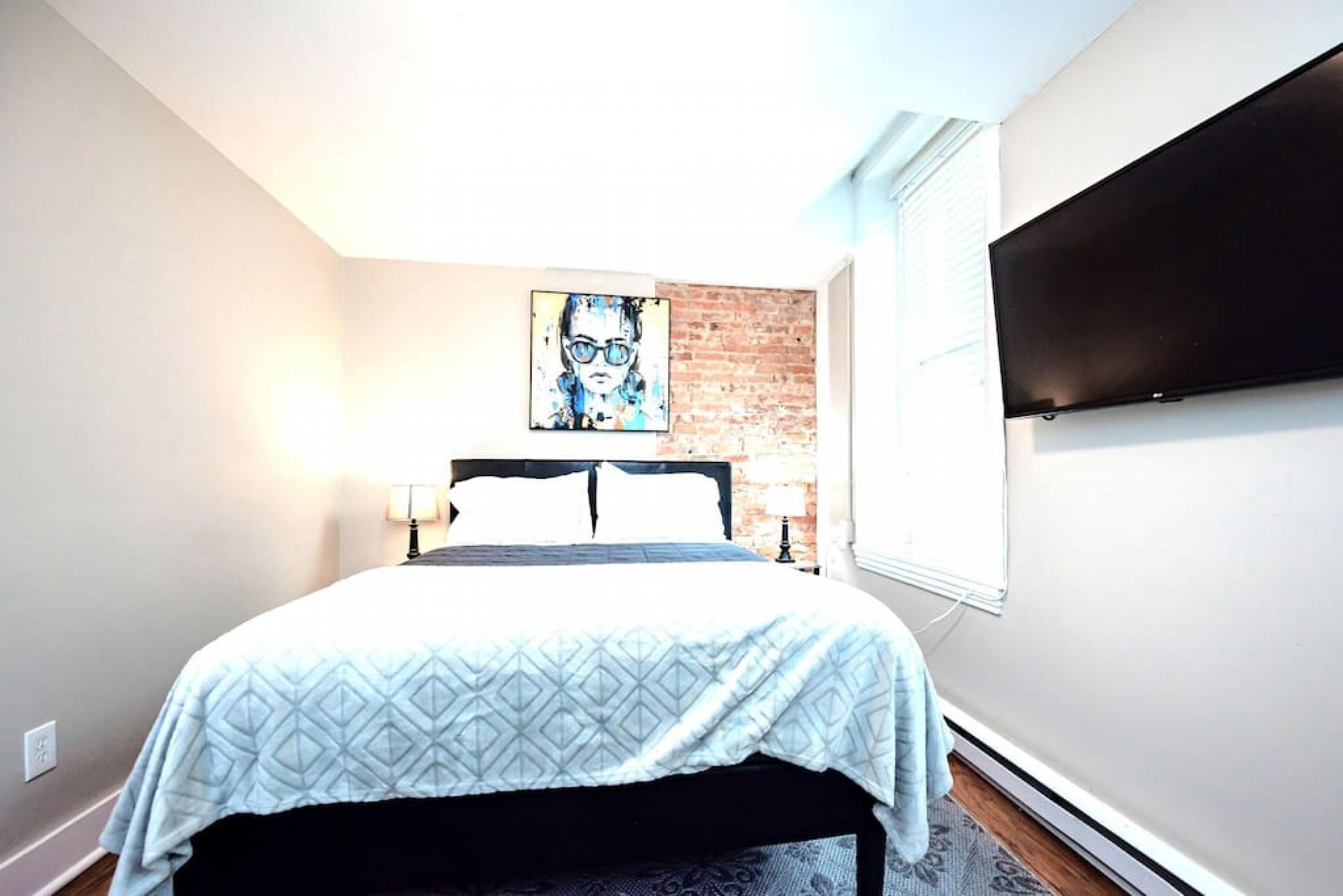 A bedroom with a bed, a brick wall, a painting, a TV, and a window.