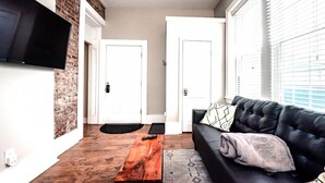 A living room with a black leather sofa, a wooden coffee table, and a flat-screen TV mounted on a brick wall. - Park Place- Cozy Modern Space Fully Updated Fast Wifi Good Coffee and Easy Parking (Wilmington)