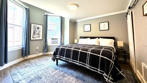 Ein Schlafzimmer mit Bett, einem Fenster mit Vorhängen, einem Heizkörper und zwei gerahmten Bildern an der Wand.