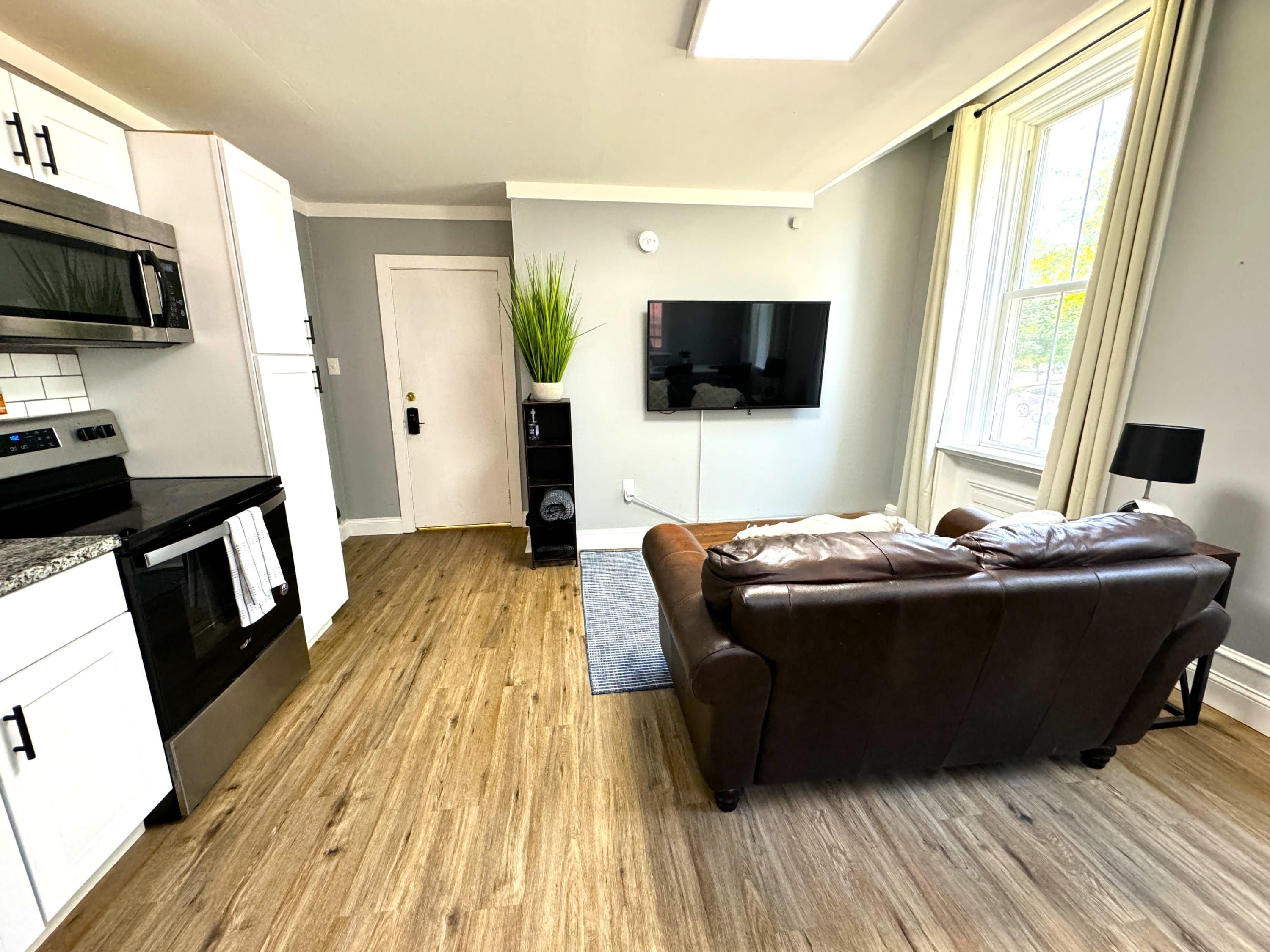 A modern kitchen with a built-in microwave and oven, a dining area with a brown leather couch, a flat-screen TV mounted on the wall, and a large window providing natural light.