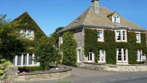A stone building with ivy-covered walls and multiple windows, surrounded by a stone wall and a small garden. - Cotteswold House (Cirencester)