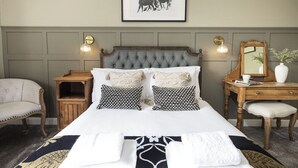 A neatly arranged bedroom with a tufted headboard, a bedside table with a lamp, a chair, and a dresser with a mirror. - Cotteswold House (Cirencester)