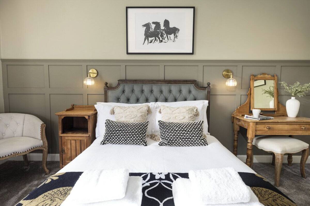 A neatly arranged bedroom with a tufted headboard, a bedside table with a lamp, a chair, and a dresser with a mirror.