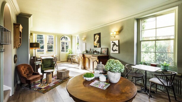 A spacious living room with a wooden dining table, comfortable seating, and a view through large windows. - Cotteswold House (Cirencester)