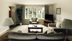 A living room with a sofa set, a coffee table, a TV stand, and a large window with curtains. - Cotteswold House (Cirencester)
