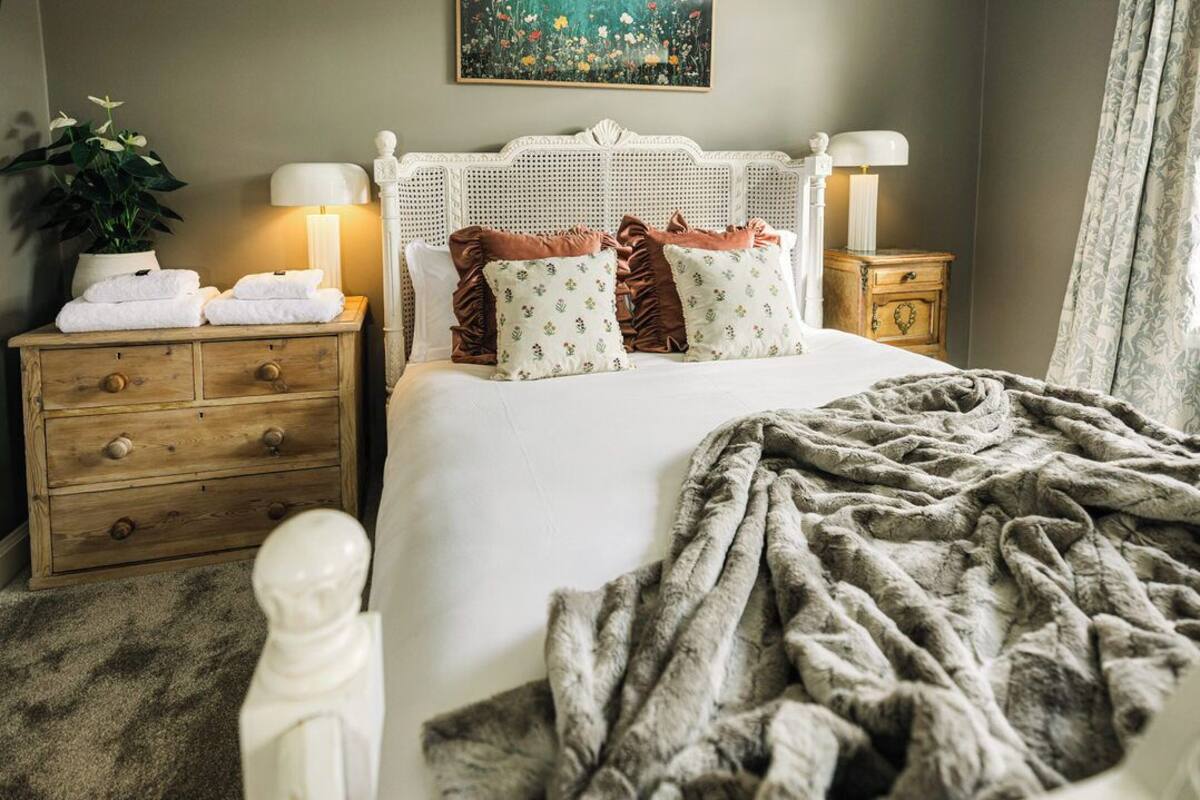 A neatly made bed with a white headboard, two bedside tables with lamps, a potted plant, and a painting on the wall.
