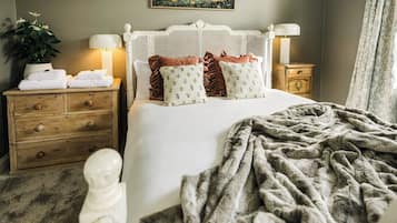 A neatly made bed with a white headboard, two bedside tables with lamps, a potted plant, and a painting on the wall.