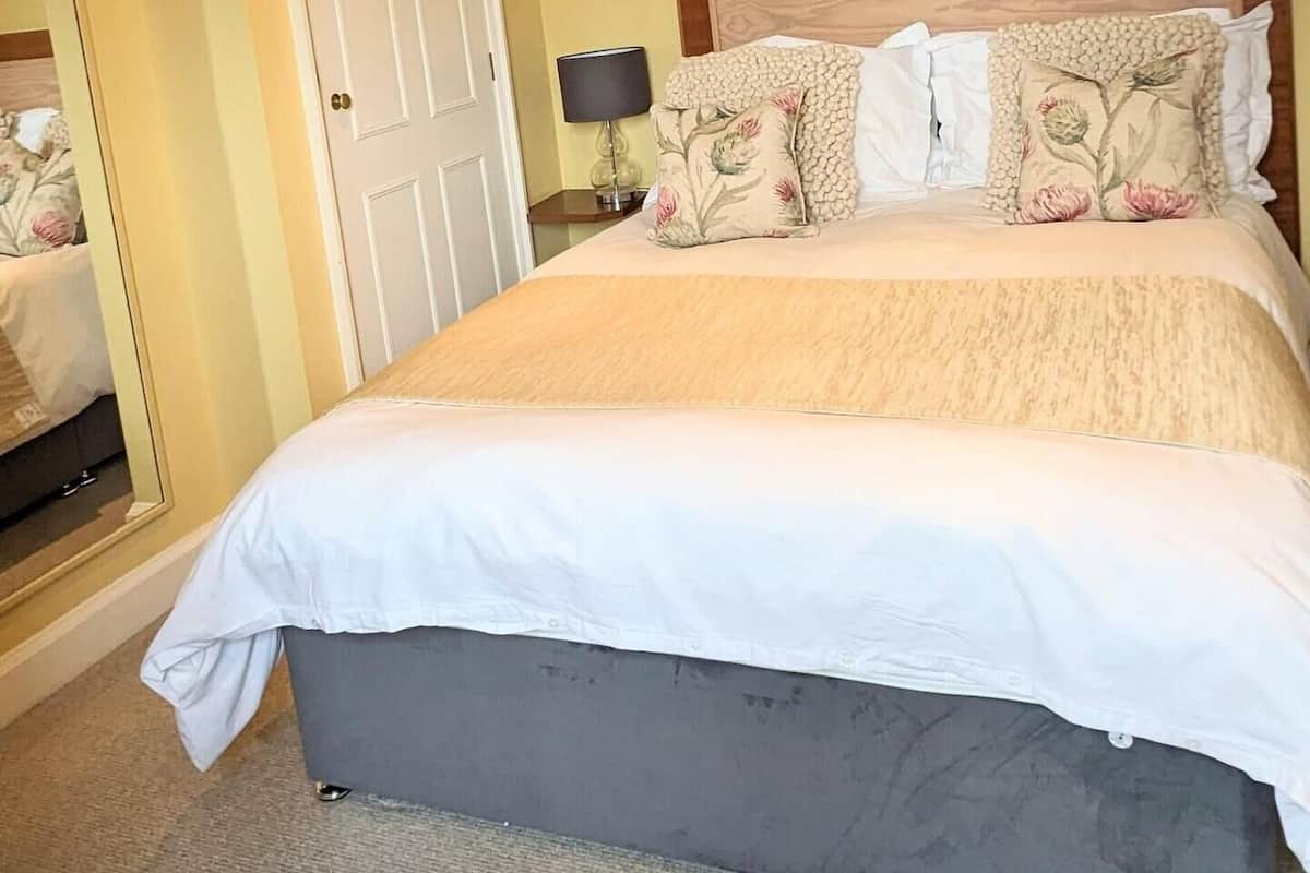 A neatly made bed with a white comforter and floral pillows, a bedside lamp, and a door with a mirror reflection.