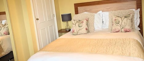 A neatly made bed with a white comforter and floral pillows, a bedside lamp, and a door with a mirror reflection.