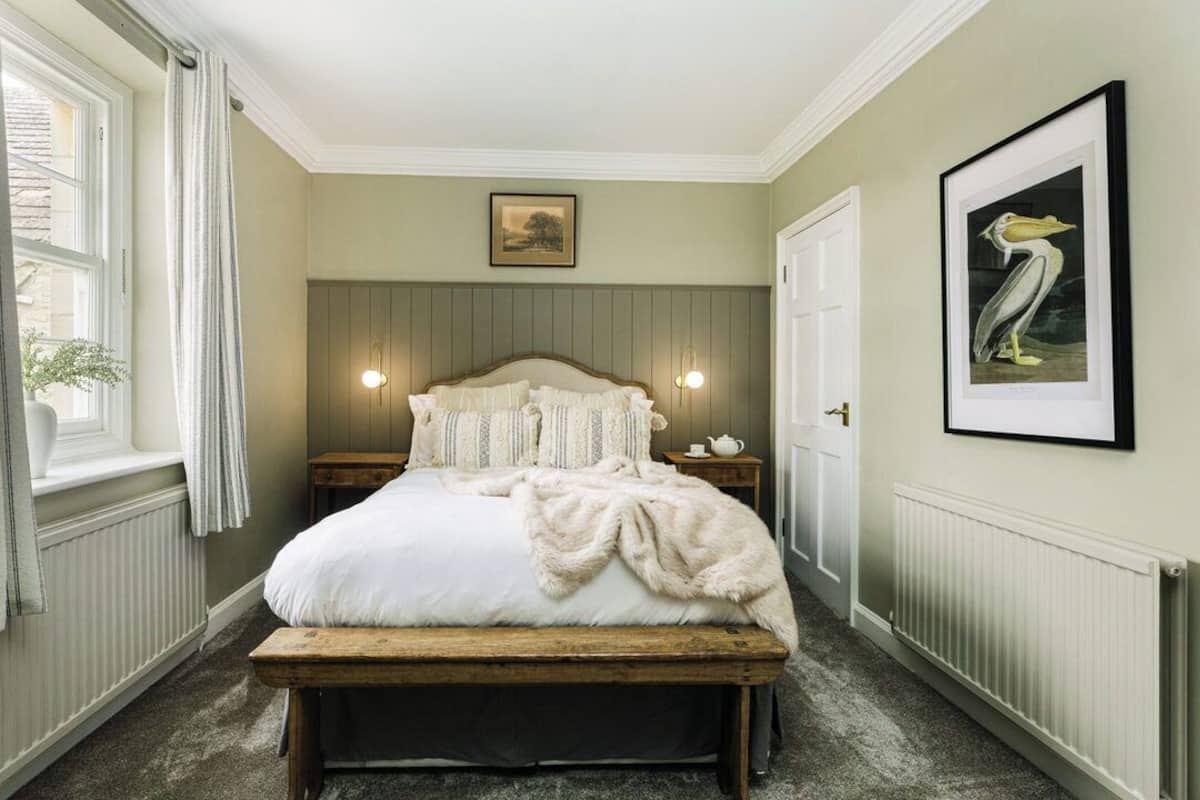 A bedroom with a bed, bedside tables, a bench, a radiator, a window with curtains, and framed artwork on the walls.