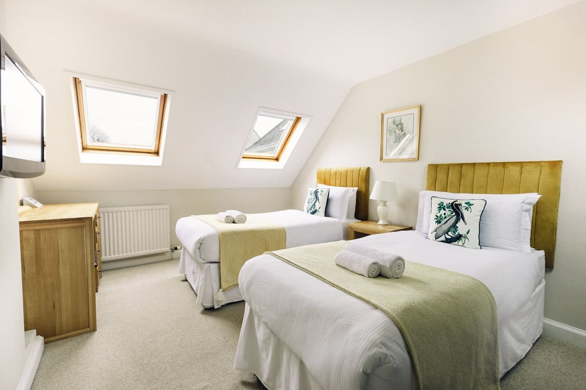 A hotel room with two beds, a desk, and a radiator. There are skylights and a painting on the wall.