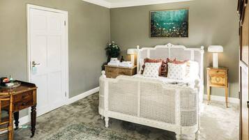 A bedroom with a white rattan bed, a wooden nightstand, a bedside table with a lamp, a door, and a painting on the wall.